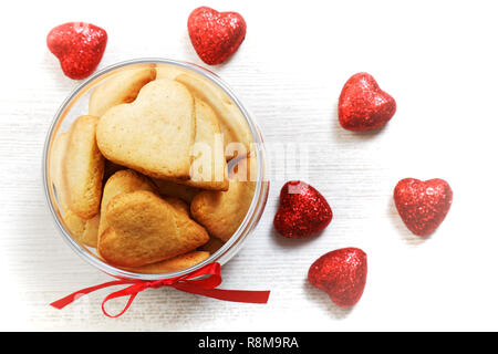 In casa a forma di cuore i cookie in un vasetto di vetro e Cuori rossi piccoli per il giorno di San Valentino in bianco sullo sfondo di legno. Vista dall'alto. Foto Stock