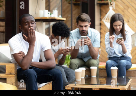 Annoiato americano africano uomo seduto con persone con telefono dipendono Foto Stock