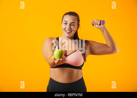 Ritratto di bella donna sovrappeso nel reggiseno sportivo azienda apple e il sollevamento di pesi isolate su sfondo giallo Foto Stock