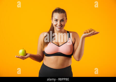 Immagine della comunità chubby donna in tuta azienda apple e croissant in entrambe le mani isolate su sfondo giallo Foto Stock