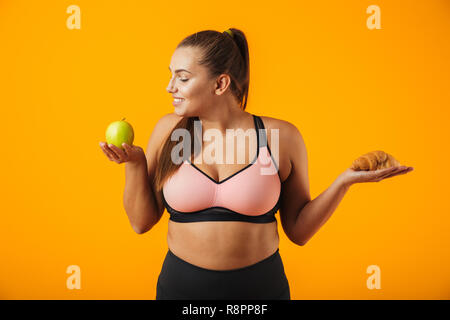 Immagine della giovane donna paffuto in tuta azienda apple e croissant in entrambe le mani isolate su sfondo giallo Foto Stock