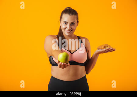 Immagine di una sana chubby donna in tuta azienda apple e croissant in entrambe le mani isolate su sfondo giallo Foto Stock