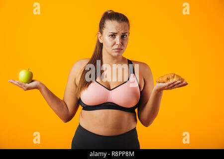 Immagine di perplesso chubby donna in tuta azienda apple e croissant in entrambe le mani isolate su sfondo giallo Foto Stock