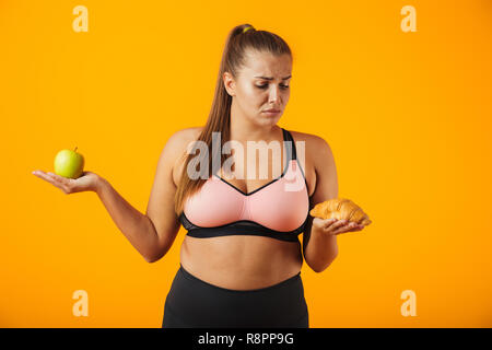 Immagine di orinato chubby donna in tuta azienda apple e croissant in entrambe le mani isolate su sfondo giallo Foto Stock