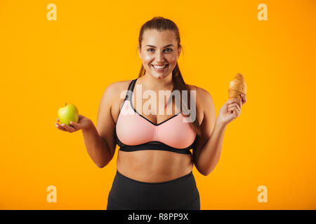 Immagine di sorridere chubby donna in tuta azienda apple e croissant in entrambe le mani isolate su sfondo giallo Foto Stock