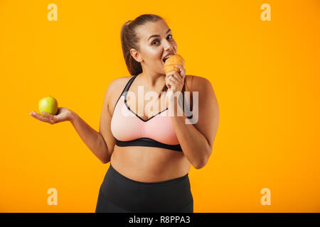 Immagine di pretty chubby donna in tuta azienda apple e mangiare croissant isolate su sfondo giallo Foto Stock