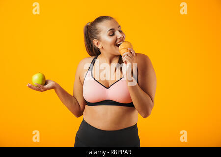Immagine di adorabili chubby donna in tuta azienda apple e mangiare croissant isolate su sfondo giallo Foto Stock