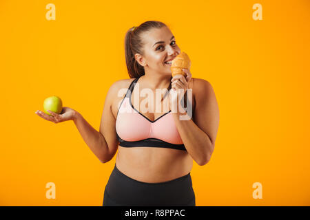 Immagine di soggetti di razza caucasica chubby donna in tuta azienda apple e mangiare croissant isolate su sfondo giallo Foto Stock