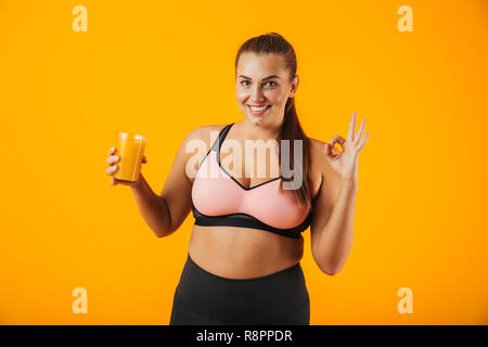 Immagine di una sana chubby donna in tuta sorridente e tenendo il vetro con succo di arancia isolate su sfondo giallo Foto Stock