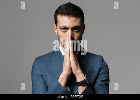 Immagine di unshaved imprenditore arabo 30s in abito formale holding palms insieme per pregare isolate su uno sfondo grigio Foto Stock