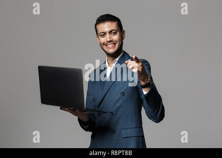 Immagine di bello imprenditore arabo 30s in abito formale sorridente e azienda laptop isolate su uno sfondo grigio Foto Stock