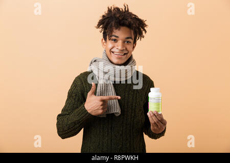 Ritratto di un felice del giovane africano vestito in abiti d'autunno isolate su sfondo beige, illustrante il contenitore con le pillole, puntamento Foto Stock