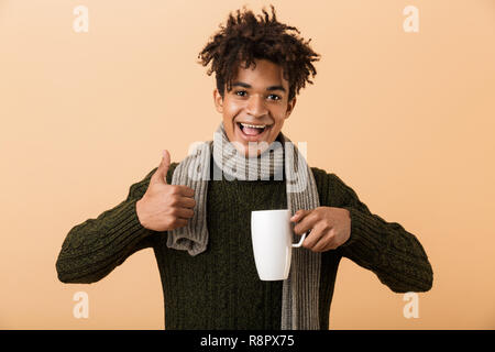 Close up ritratto di un felice del giovane africano vestito in abiti d'autunno isolate su sfondo beige, tenendo una tazza, mostrando pollice in alto Foto Stock