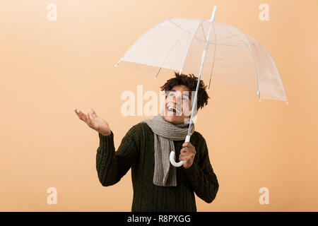 Ritratto di entusiasti african american ragazzo indossa un maglione e sciarpa in piedi sotto ombrellone isolate su sfondo beige Foto Stock