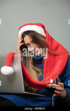 Ragazza solitaria in un cappello di Natale è bere vino, lavorando sul computer portatile Foto Stock