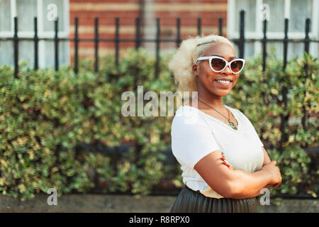 Ritratto sorridente, fiducioso giovane donna in occhiali da sole sul marciapiede urbano Foto Stock