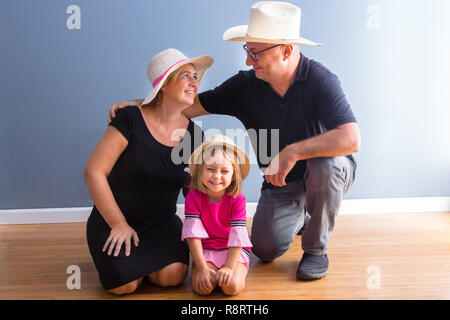 Donna di mezza età e genitori man in black casual sono in basso verso il pavimento della camera accanto alla loro giovane figlia in abito rosa, indossare un cappello di paglia Foto Stock