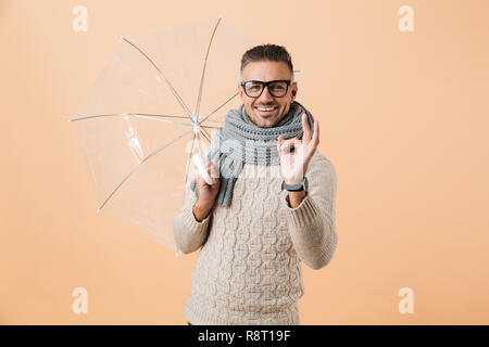 Ritratto di un allegro uomo vestito di maglione e sciarpa in piedi sotto ombrellone isolate su sfondo beige, mostrando ok Foto Stock