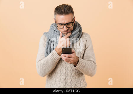 Ritratto di un pensieroso uomo vestito di maglione e sciarpa in piedi isolato su sfondo beige, utilizzando il telefono cellulare Foto Stock