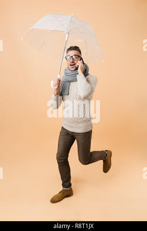 A piena lunghezza verticale se un bel uomo vestito di maglione e sciarpa camminare sotto ombrellone isolate su sfondo beige, parlando al telefono cellulare Foto Stock