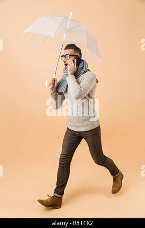 A piena lunghezza verticale se sorpreso un uomo vestito con un maglione e sciarpa camminare sotto ombrellone isolate su sfondo beige, parlando al telefono cellulare Foto Stock