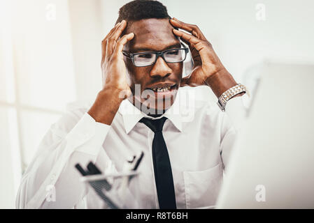 Ansioso imprenditore africano si trova in ufficio e mantiene la testa con le mani. Foto Stock