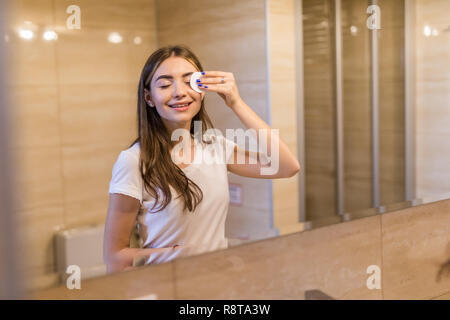 Focalizzato bella giovane donna pulizia la sua pelle con un tampone di cotone, guardando lo specchio nel bagno di casa. Bellezza e cura della pelle concetto, lo stile di vita Foto Stock