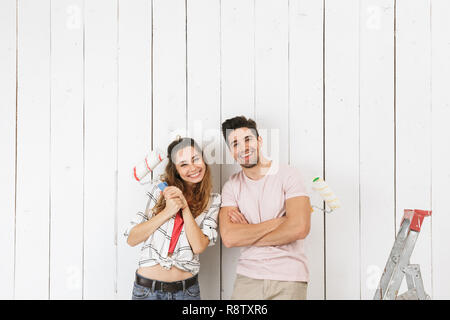 Foto di sorridere giovane uomo e donna pittura muro bianco e facendo lavori di rinnovo utilizzando rulli di vernice Foto Stock