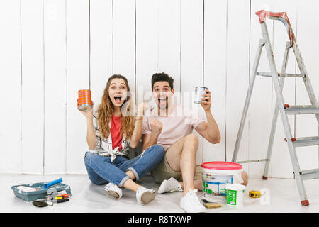 Foto di eccitato giovane uomo e donna seduta sul pavimento oltre il muro bianco vicino alla scaletta e azienda barattoli di vernice mentre effettua lavori di ristrutturazione interni Foto Stock