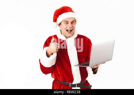 Ritratto di un allegro giovane uomo vestito di santa claus costume isolato permanente su sfondo bianco, tenendo il computer portatile, mostrando pollice in alto Foto Stock