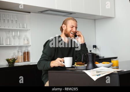Bel giovane avente la colazione leggendo il giornale mentre è seduto alla cucina, parlando al telefono cellulare Foto Stock