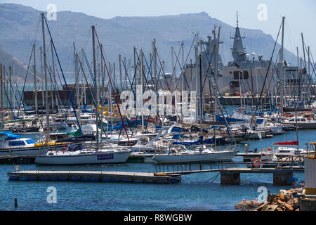 Yacht ormeggiati nel porto o porto di Simonstown in Città del Capo su un soleggiato ma ventoso giorno d'estate con un mezzo navale o fregata in background Foto Stock