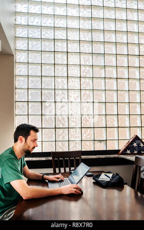 Giovane uomo barbuto in casa a lavorare in remoto sul computer portatile in parte anteriore del blocco di vetro parete Foto Stock