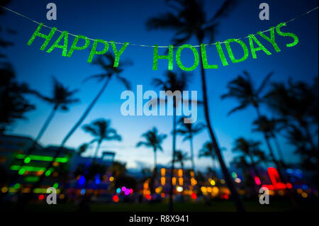 Vacanze felici messaggio appesi in glittery banner di partito al di sopra di alberi di palma tropicali dell'incandescente luci al neon della skyline di Miami Beach, Florida Foto Stock