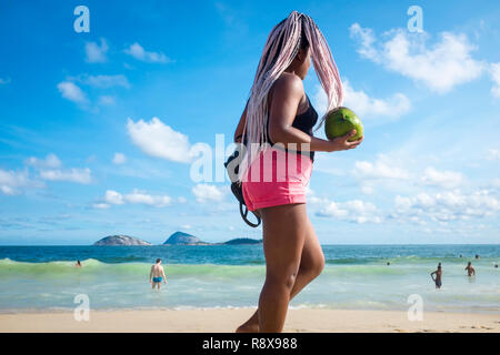 RIO DE JANEIRO - Gennaio 2018: una giovane donna con rosa capelli intrecciati passeggiate con un fresco noce di cocco verde sulla riva della spiaggia di Ipanema. Foto Stock