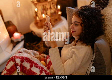 Bella giovane donna con capelli ricci è la lettura dei messaggi sul suo smartphone con decorazione di Natale in background Foto Stock