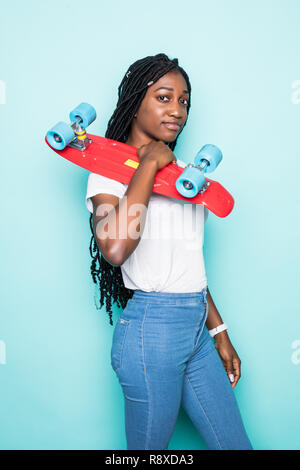 Straordinaria giovane donna in jeans e maglietta rosa indossando occhiali da sole in posa con sorriso sincero su sfondo blu. Carino ragazza africana holding e skateboard Foto Stock