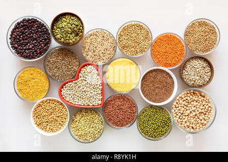 Vista dall'alto di ciotole in vetro con cereali, fagioli e semi con cuore rosso a forma di ciotola in medio su sfondo bianco. Foto Stock