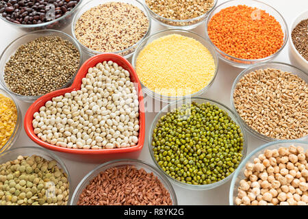Close-up ciotole in vetro con cereali, fagioli e semi con cuore rosso a forma di ciotola in medio su sfondo bianco. Foto Stock