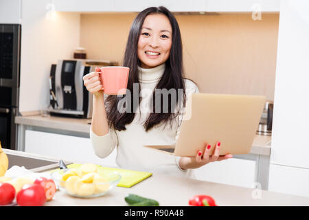 Immagine dei giovani brunette con la coppa in mano al tavolo con i laptop in cucina Foto Stock