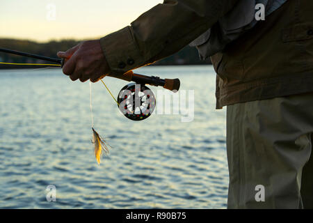 Un primo piano delle mani di un pescatore a mosca tenendo la sua canna da mosca e l'aspo e un "Lefty's Deceiver" streamer fly. Foto Stock