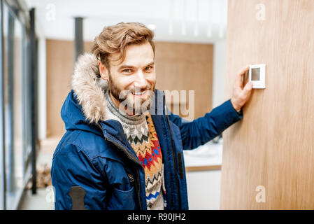 Uomo in abbigliamento invernale sensazione di freddo in camera di regolazione di temperatura con termostato elettronico a casa Foto Stock