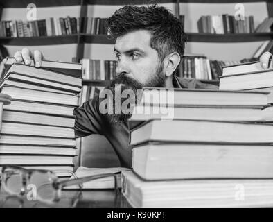 Insegnante o studente con la barba si siede a tavola con gli occhiali, sfocato. Concetto di bibliofili. Uomo sulla faccia di rigorosa tra pile di libri, mentre studiava in biblioteca, scaffali, sullo sfondo. Foto Stock