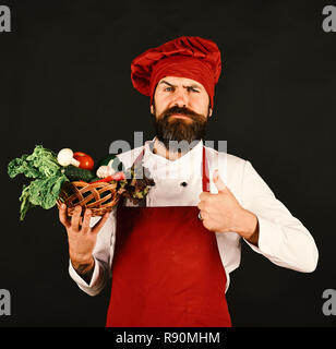 Lo Chef detiene la lattuga, pomodoro, pepe e funghi. Un sano concetto di cucina a vista. Cucinare con un rigoroso volto in Borgogna mantiene uniforme verdure nella ciotola che mostra Thumbs up. Uomo con la barba su sfondo nero. Foto Stock