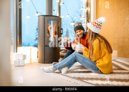 Coppia giovane vestito in bright maglioni e cappelli seduti insieme vicino al camino nella casa moderna durnig Inverno tempo Foto Stock