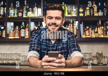 Giovane barista in piedi appoggiata sul banco bar tenendo lo smartphone cerca fotocamera a sorridere allegro vista frontale Foto Stock