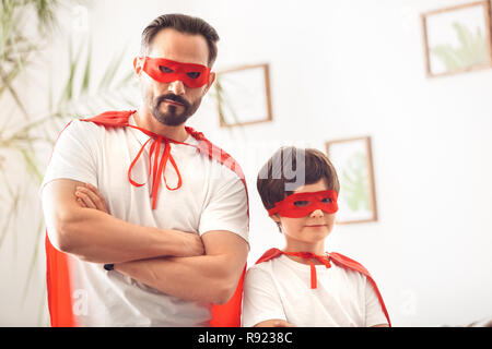 Padre e figlio indossando costumi superheroe insieme a casa in piedi che guarda la fotocamera in maschere bracci incrociati gravi Foto Stock