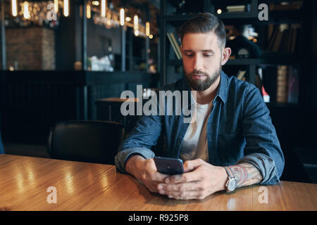 Giovane imprenditore barbuto,vestito con una camicia in denim, seduti a tavola in cafe e utilizza lo smartphone. Uomo che utilizza gadget. Foto Stock