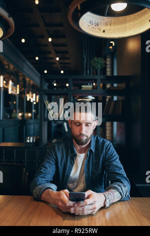 Giovane imprenditore barbuto,vestito con una camicia in denim, seduti a tavola in cafe e utilizza lo smartphone. Uomo che utilizza gadget. Foto Stock