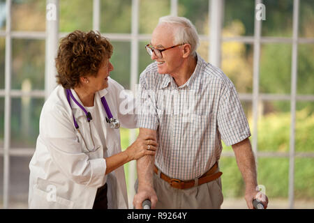Donna sorridente medico incoraggiando l'anziana paziente di sesso maschile che sta usando il suo telaio a piedi. Foto Stock
